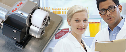 links ein Schlauchstripper, rechts eine Frau und ein Mann in einem Labor, im Hintergrund ist eine Mulltisafe Entsorgungsbox zu sehen, daneben Verpackungen der S-Monovette, darüber drei Probenständer gefüllt mit Monovetten